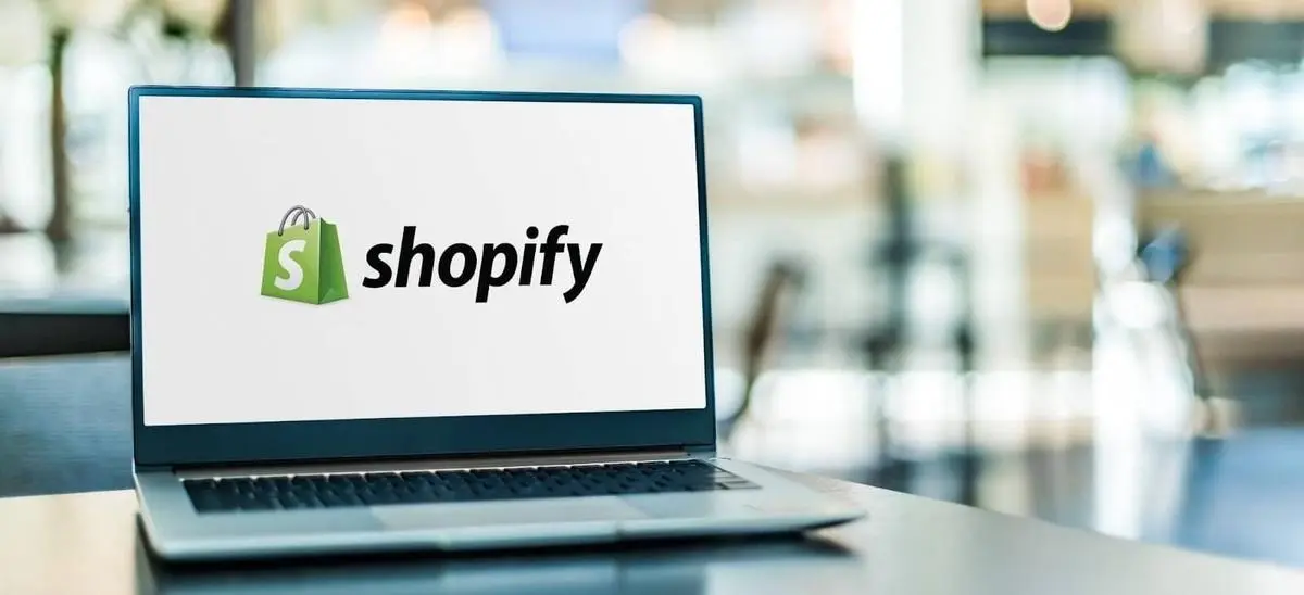 Ein Tisch im Vordergrund auf dem ein aufgeklappter Laptop mit dem Shopify-Logo als Bildschirmschoner steht. Im Hintergrund sind verschwommen weitere Tische und Stühle erkennbar. Shopify mit Amazon zu verbinden bietet Onlinehändlern einige Vorteile. Welche das sind, erfährst du in diesem Beitrag!