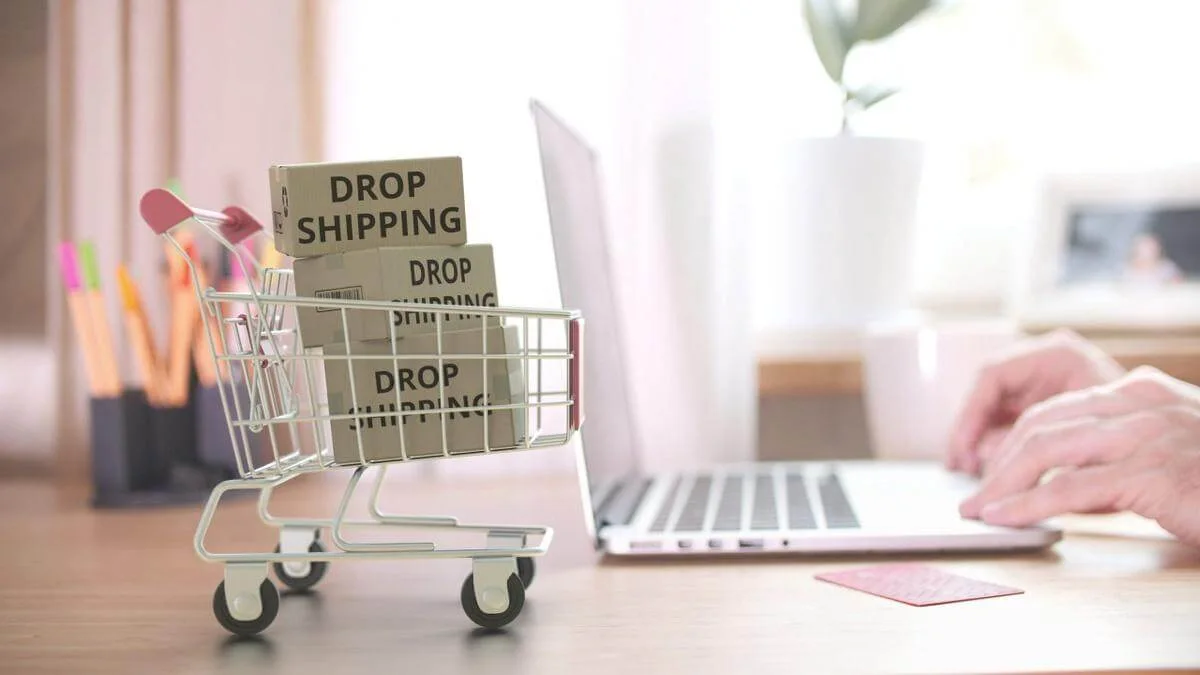 Blick von der Seite auf einen Schreibtisch auf dem im Hintergrund ein Laptop steht, an dem eine Person arbeitet. Im Vordergrund ist ein Miniatur-Einkaufswagen mit drei Paketen mit der Aufschrift "Dropshipping" zu sehen. Wir zeigen dir, worauf es beim Dropshipping ankommt! 