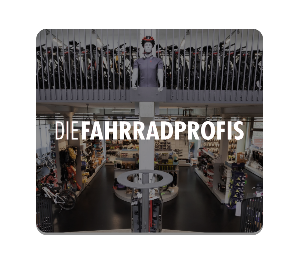 Xentral ERP for bike retail: High-angle interior view of a large two-story bicycle shop with the white text overlay "DIE FAHRRADPROFIS". The ground floor features retail displays, while the upper level shows a long row of bikes.