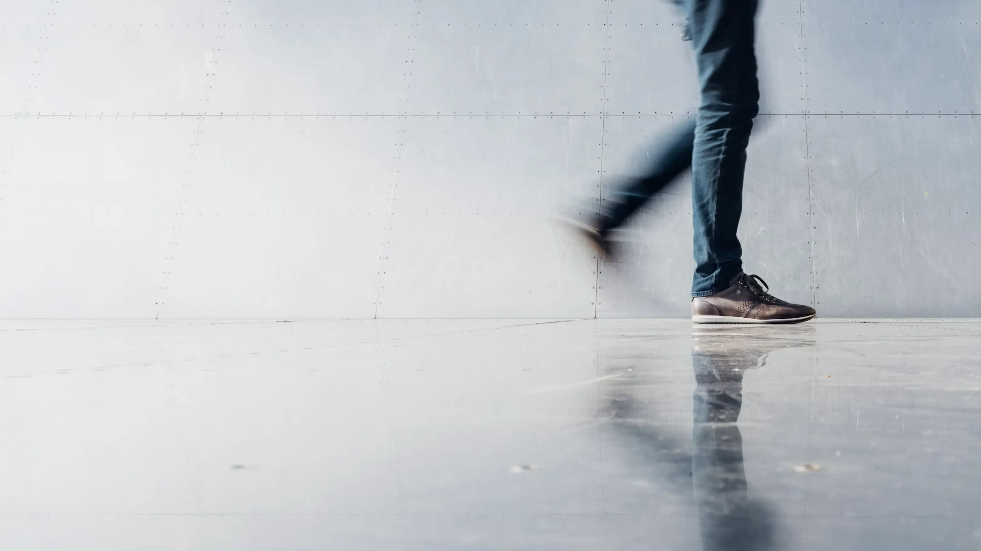 Person in Jeans und braunen Sneakern geht zügig an einer grauen Metallwand vorbei, Bewegungsunschärfe und Spiegelung auf dem Boden