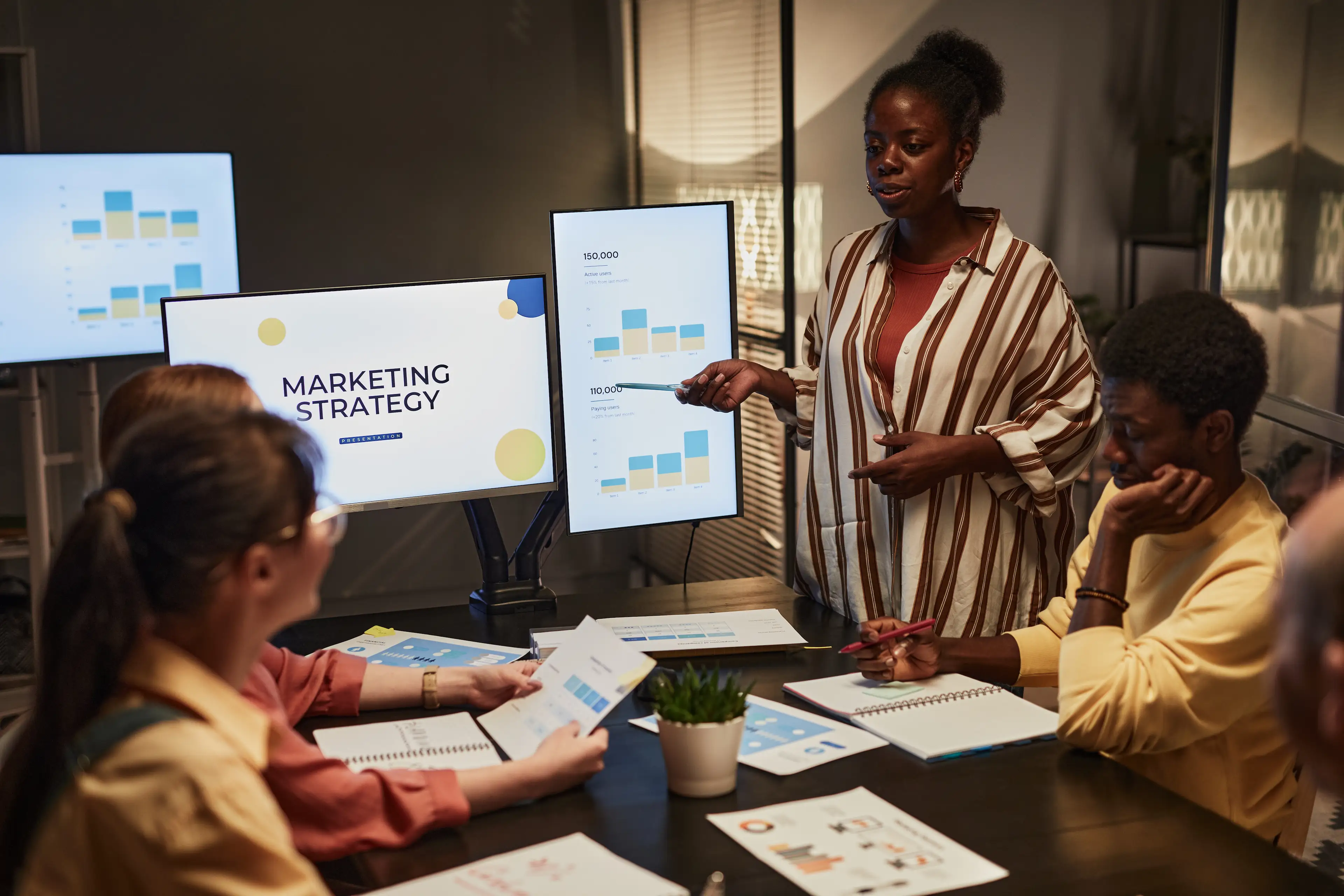 Marketingteam in einem abendlichen Meeting: Eine Frau im gestreiften Hemd präsentiert Nutzerzahlen und Balkendiagramme auf zwei Monitoren, während drei Kolleg:innen am Konferenztisch mit ausgedruckten Charts und Notizbüchern zuhören.