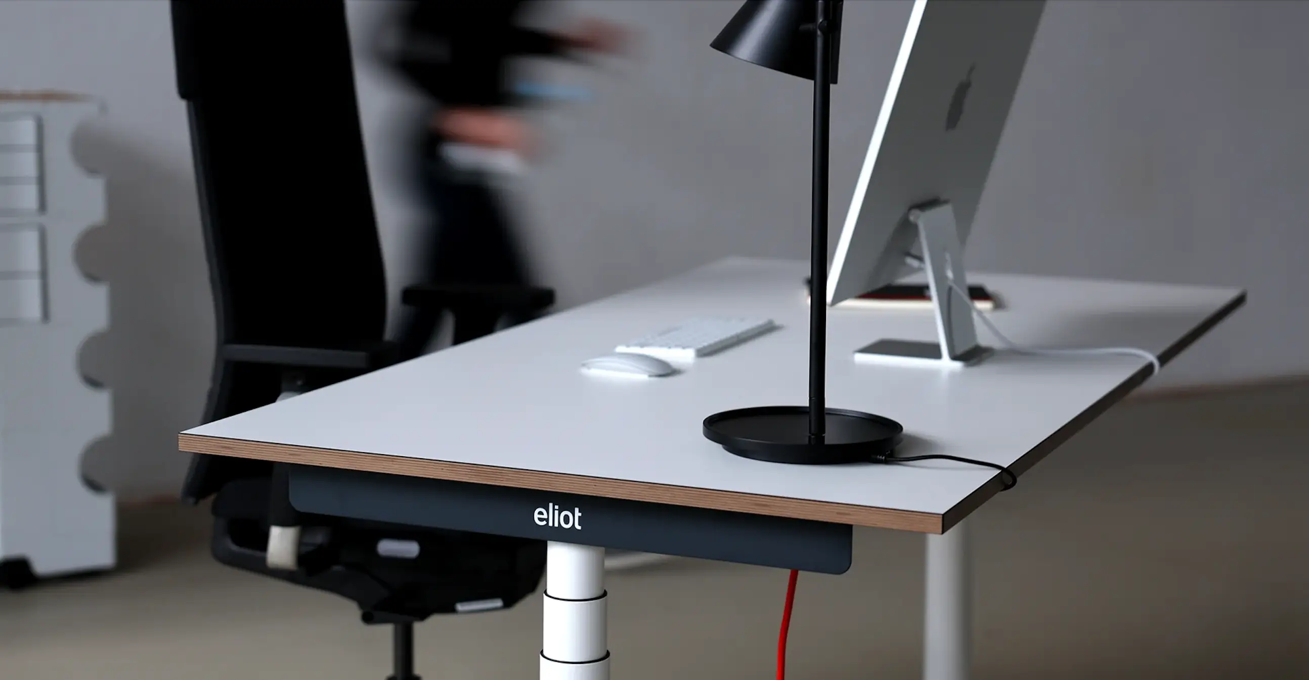 Höhenverstellbarer Eliot-Schreibtisch mit weißer Tischplatte, darauf ein iMac, Apple-Tastatur und -Maus sowie eine schwarze Tischlampe. Im unscharfen Hintergrund ein ergonomischer Bürostuhl und eine vorbeigehende Person.