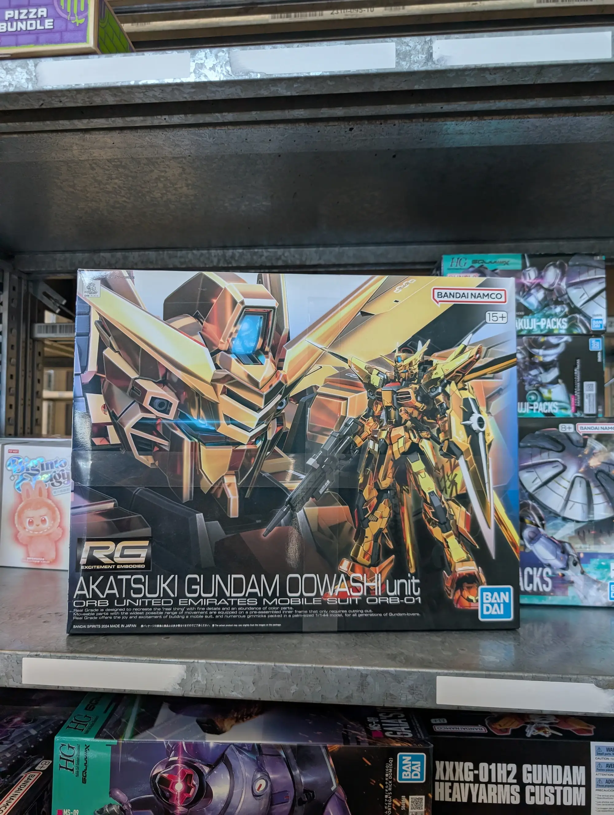 Bandai-Modellbausatz "RG Akatsuki Gundam Oowashi unit" (Maßstab 1/144) im Regal eines Hobbyshops, umgeben von weiteren Gunpla-Boxen wie HG Gundam Heavyarms Custom.