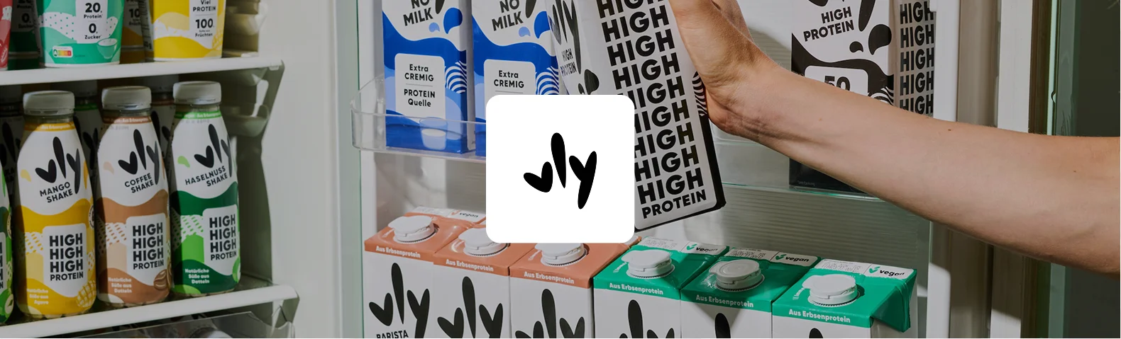 Kühlschrank gefüllt mit vly-Produkten aus Erbsenprotein: High-Protein-Shakes in Mango, Coffee und Haselnuss sowie Tetra Paks der Milchalternative "No Milk" in Varianten wie Extra Cremig, High Protein und Barista. Eine Hand greift nach einer Packung, das vly-Logo steht in der Mitte.