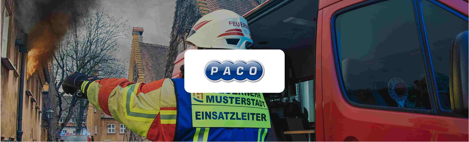 Feuerwehr-Einsatzleiter in Schutzkleidung mit Weste "Feuerwehr Musterstadt – Einsatzleiter" zeigt neben einem roten Einsatzfahrzeug auf ein brennendes Gebäude mit Rauchentwicklung. In der Bildmitte das PACO-Logo.
