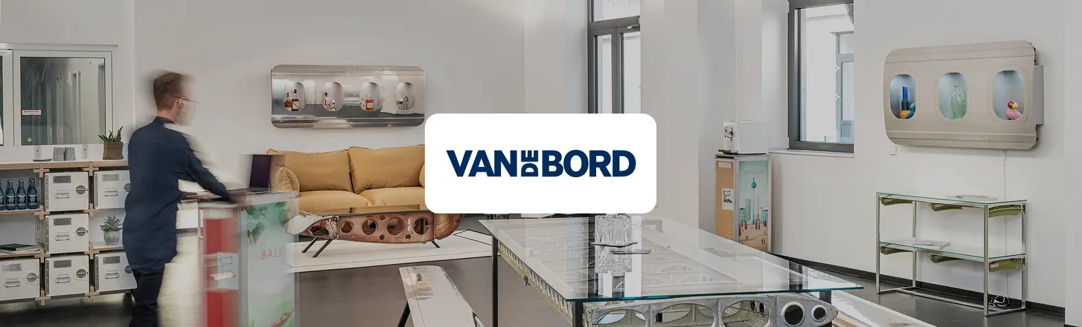 Showroom von Van de Bord mit Upcycling-Möbeln aus Flugzeugteilen: Glastisch auf Tragflächen-Struktur, Sofa auf Rumpfelementen, Wandregale aus Flugzeugfenstern und Regale aus Bordtrolleys. Eine Person steht an einem Trolley-Tresen, in der Mitte das Van-de-Bord-Logo.