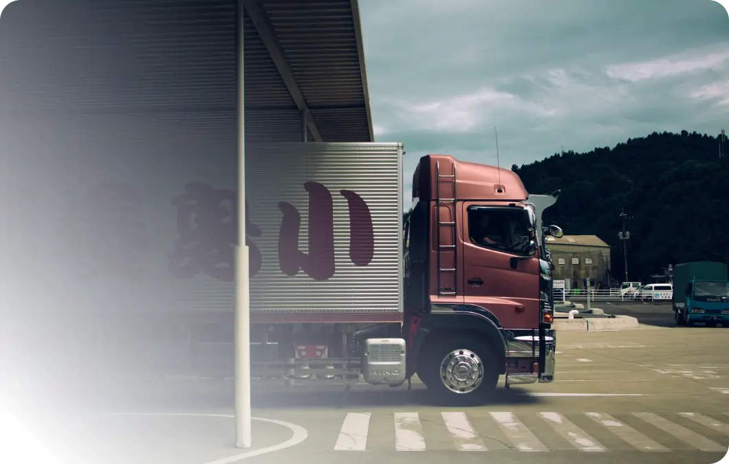 Roter Hino-Lkw mit silbernem Kofferaufbau und japanischen Schriftzeichen steht an einer Laderampe eines Logistikgebäudes, im Hintergrund bewaldete Hügel und ein weiterer Lieferwagen.