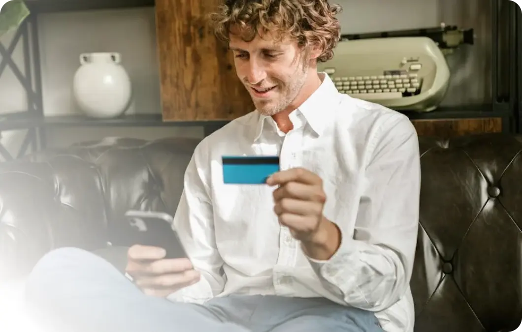 Tevreden klanten dankzij soepele checkout-processen met Xentral ERP voor e-commerce: een glimlachende man zit op een leren bank en houdt een creditcard en een smartphone in zijn handen, op de achtergrond is een typemachine op een plank te zien.